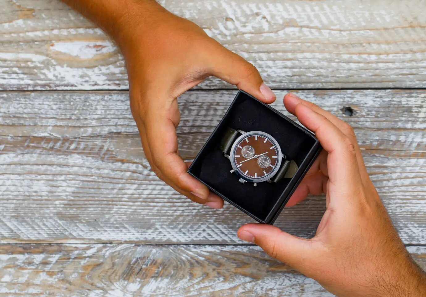 A birthday-themed flat lay on a wooden background, with hands giving and receiving a watch box.